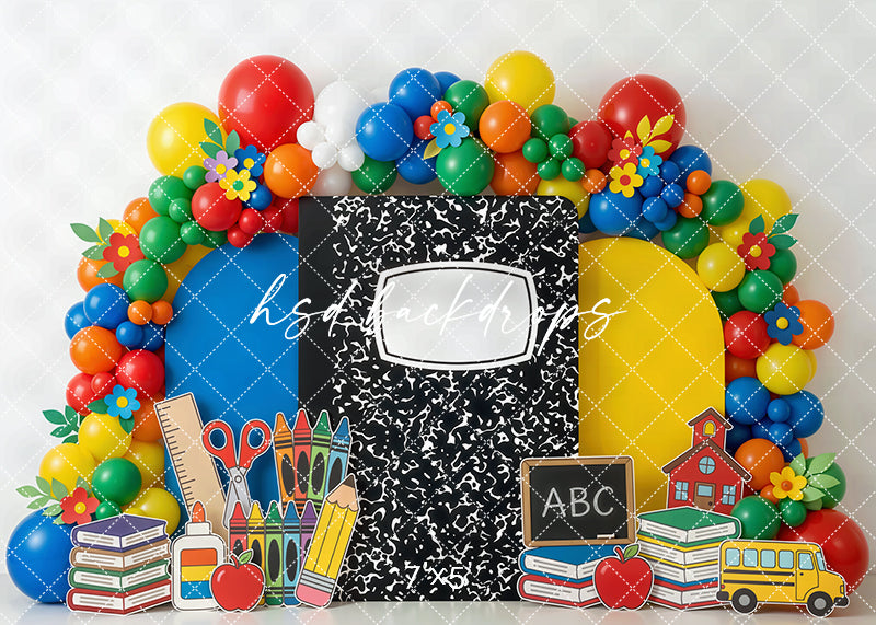 Composition notebook school photography backdrop with colorful balloon arch, books, crayons, scissors, glue, apples, chalkboard, school bus, and schoolhouse for back-to-school portraits.