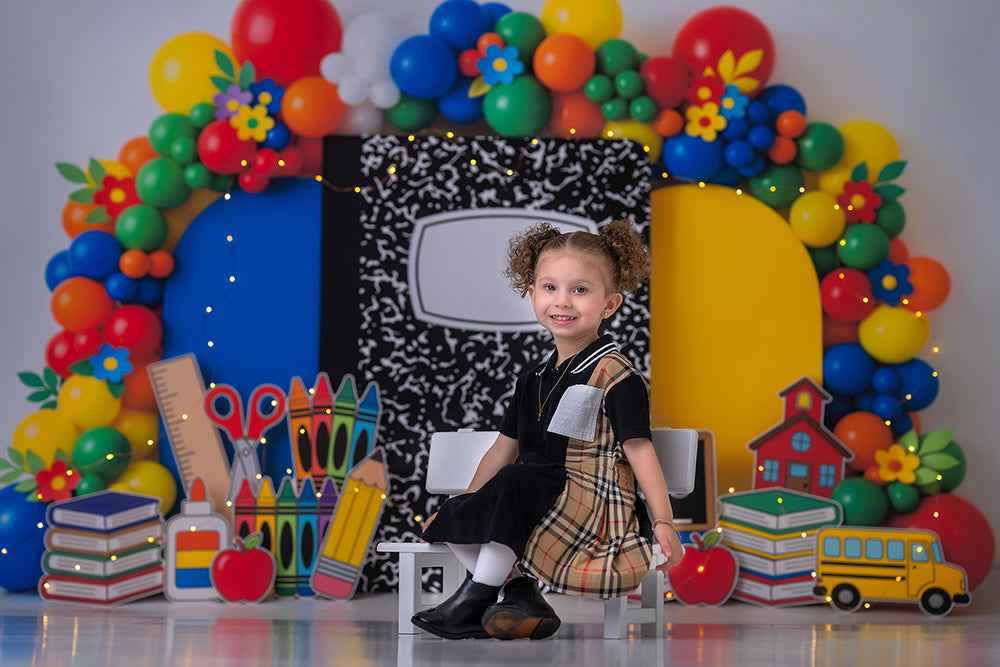 Composition Notebook School photography backdrop in use with a child seated in front of a colorful balloon arch, composition notebook centerpiece, school supply props, books, apples, and a school bus for back-to-school portraits.