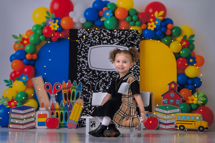 Composition Notebook School photography backdrop in use with a child seated in front of a colorful balloon arch, composition notebook centerpiece, school supply props, books, apples, and a school bus for back-to-school portraits.