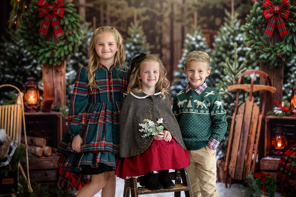 Christmas tree farm photography backdrop with snowy evergreen trees, rustic wooden posts, red plaid bows, lanterns, sled props, and cozy holiday portrait styling.