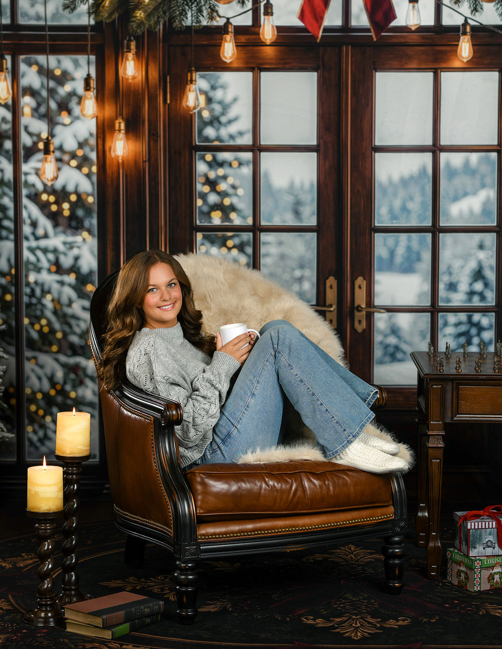 Cozy winter window Christmas photography backdrop with dark wood French doors, snowy evergreen view, warm hanging lights, holiday garland, candles, leather chair, and classic winter portrait styling.