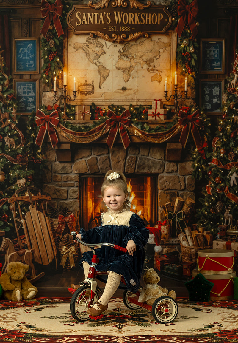 Santa’s Workshop fireplace photography backdrop with a glowing stone fireplace, North Pole map, Christmas garland, red bows, toys, sled, teddy bears, and cozy holiday workshop styling.
