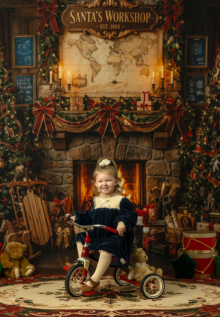 Santa’s Workshop fireplace photography backdrop with a glowing stone fireplace, North Pole map, Christmas garland, red bows, toys, sled, teddy bears, and cozy holiday workshop styling.