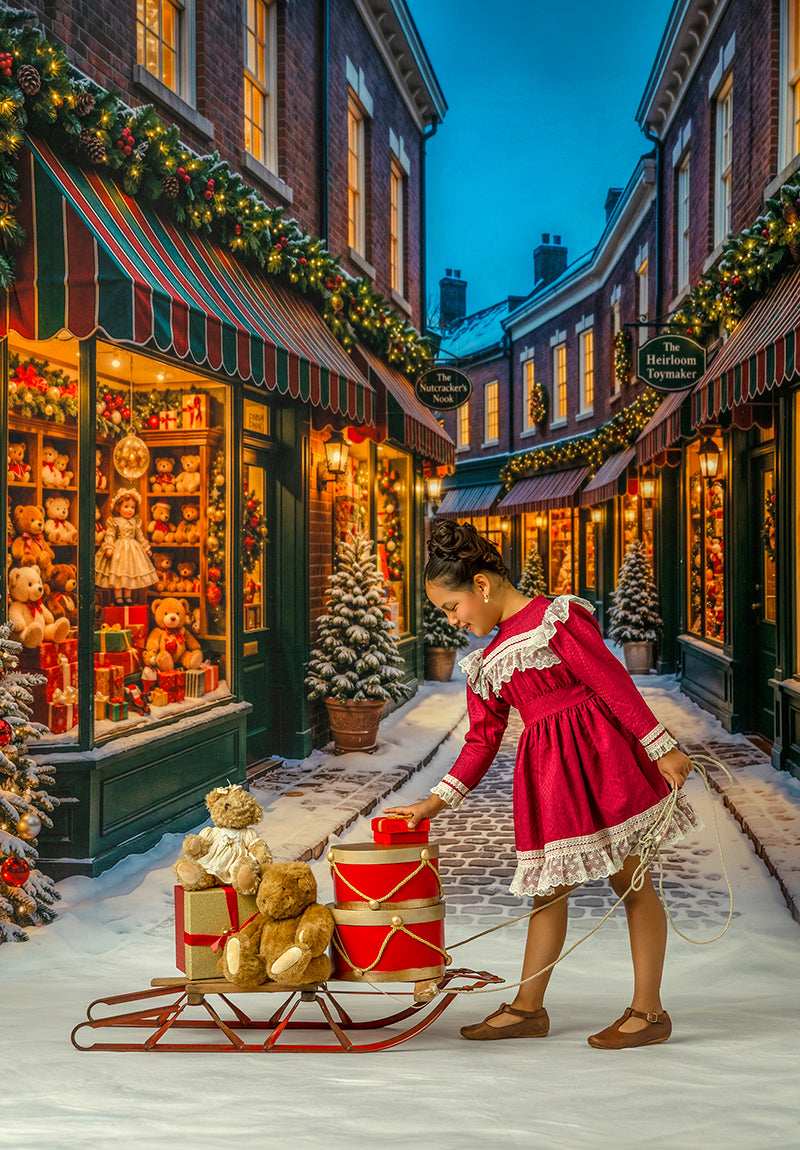 Christmas toy shop street photography backdrop with snowy village storefronts, glowing holiday windows, decorated trees, teddy bears, gifts, sleigh prop, and festive Christmas portrait styling.