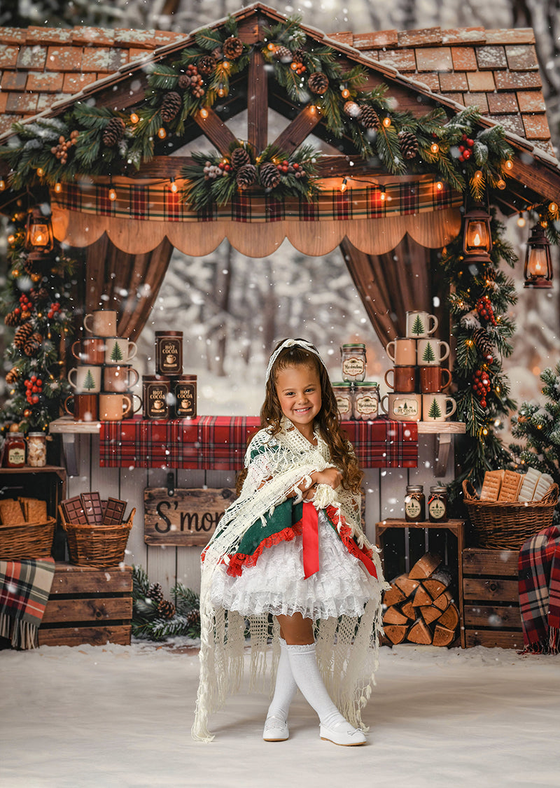 Christmas hot cocoa stand photography backdrop with rustic wooden cocoa booth, plaid accents, evergreen garland, lanterns, mugs, s’mores, snowy trees, and cozy holiday styling.