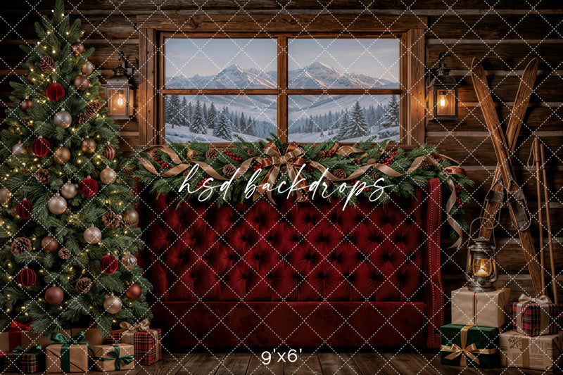 Cozy Cabin Christmas photography backdrop with rustic wood cabin walls, snowy mountain window view, burgundy tufted sofa, decorated Christmas tree, evergreen garland, lanterns, skis, wrapped gifts, and warm holiday styling.