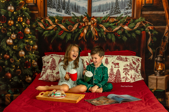 Cozy holiday cabin bed backdrop with red bedding, rustic wood walls, snowy pine window view, decorated Christmas tree, warm lantern light, and classic Christmas portrait styling.