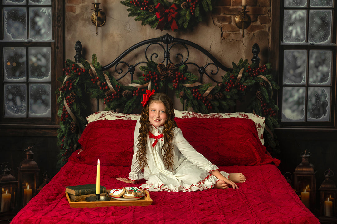 Evergreen Bedframe Christmas photography backdrop with black iron bedframe, red bedding, snowy windows, rustic brick wall, evergreen garland, red berries, pinecones, candles, and cozy holiday styling.