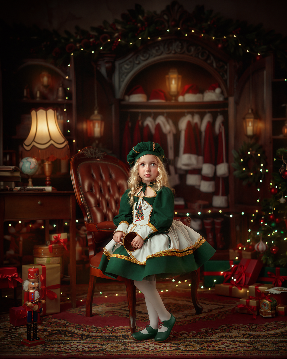 Vintage Santa's Closet Christmas portrait photographed by MK Slowinski featuring a young girl in a forest green holiday dress seated on an antique leather chair, surrounded by Santa coats, glowing lanterns, evergreen garlands, and wrapped Christmas gifts.