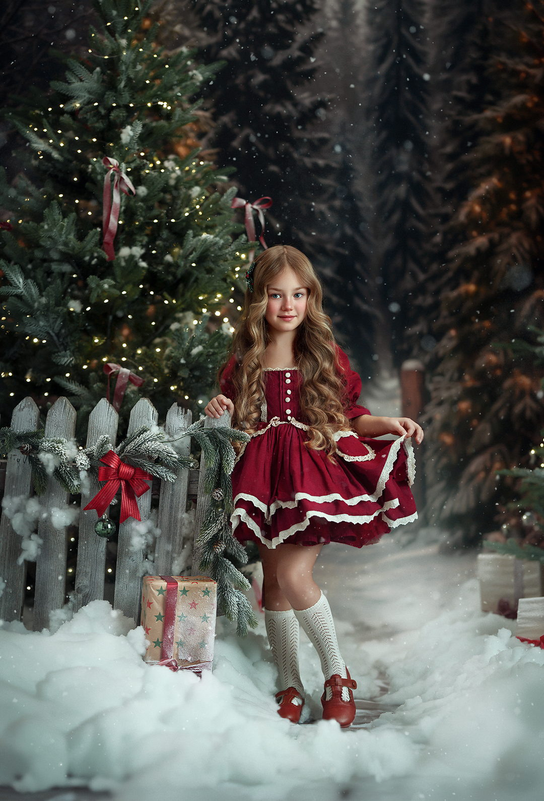 Rustic snowy Christmas tree photography backdrop captured by MK Slowinski with a child in a burgundy Christmas dress, decorated evergreen tree, wooden fence, and falling snow.
