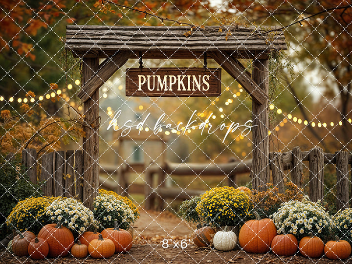 Rustic fall photography backdrop featuring a wooden archway with a "Pumpkins" sign, surrounded by orange pumpkins, white and yellow mums, string lights, and autumn foliage.