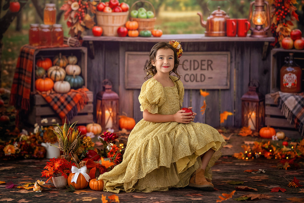 Child posing in front of an autumn apple cider stand photography backdrop with rustic wood booth, apple baskets, fall leaves, pumpkins, lanterns, plaid accents, and warm harvest styling.