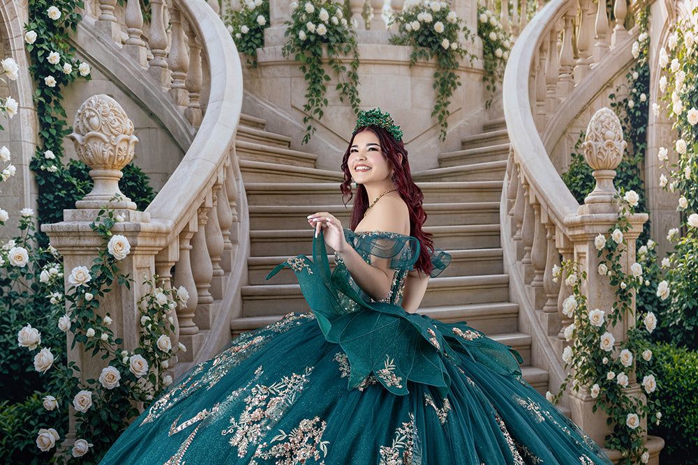 Elegant quinceañera photography backdrop featuring a grand ivory palace staircase adorned with white roses and greenery, with a quinceañera wearing an emerald green ball gown and jeweled crown.