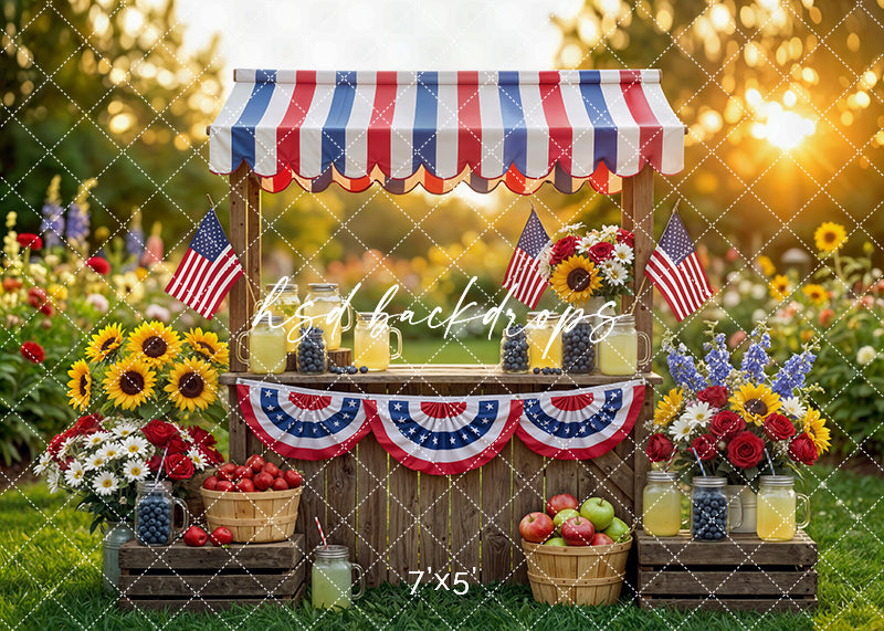 Patriotic lemonade stand photography backdrop with American flags, red white and blue decor, fresh fruit, and summer floral garden for Fourth of July portraits.