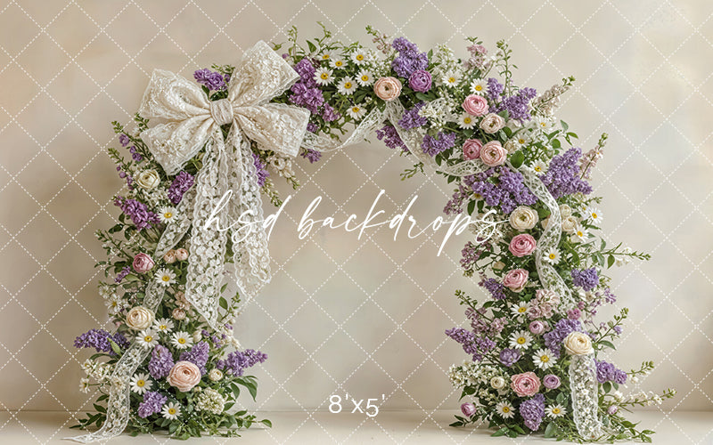 Lavender and blush floral arch backdrop with lace bow, lilacs, roses, daisies, and soft spring garden details