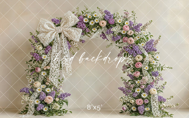 Lavender and blush floral arch backdrop with lace bow, lilacs, roses, daisies, and soft spring garden details