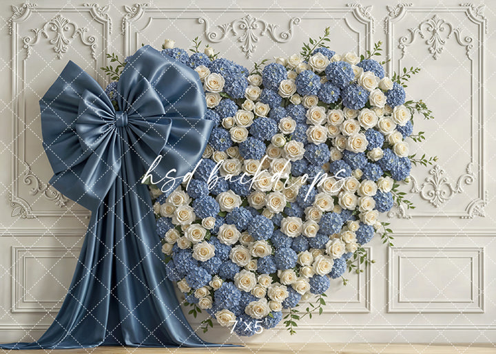 Dusty blue hydrangea and ivory rose heart floral wall with large satin bow against ornate white paneling for elegant photography sessions.