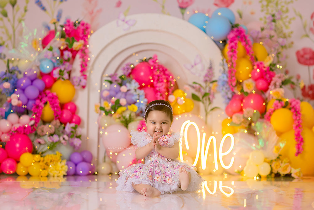 One Wildflower cake smash photography backdrop with layered white arches, colorful balloons, bright florals, butterflies, and a glowing one sign for girls birthday portraits.