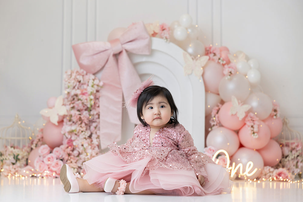 Pink Butterfly Bow cake smash photography backdrop with a white arch, blush and ivory balloons, oversized pink bow, florals, butterflies, and glowing one sign for girls birthday portraits.