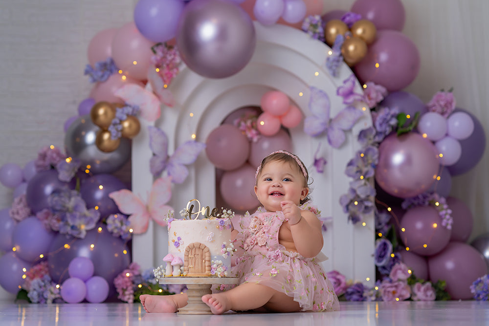 Lavender butterfly garden photography backdrop with a white layered arch, pastel purple and pink balloon garlands, floral accents, and butterflies for whimsical birthday portraits.