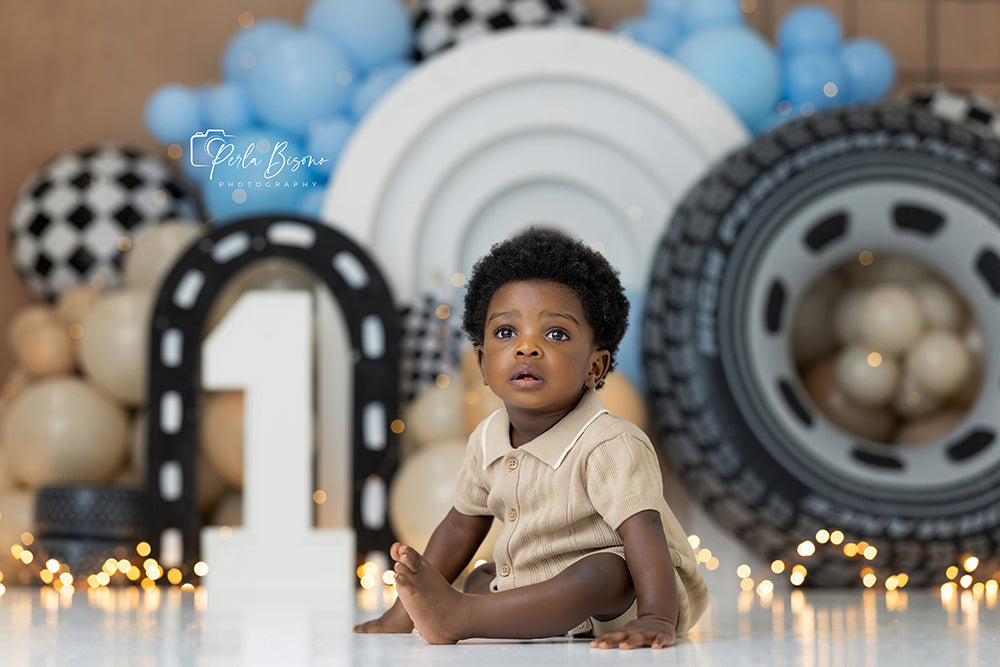 Victory Lap race car photography backdrop with blue balloon garland, oversized tire, checkered flag accents, road arches, trophy details, and birthday cake smash styling.