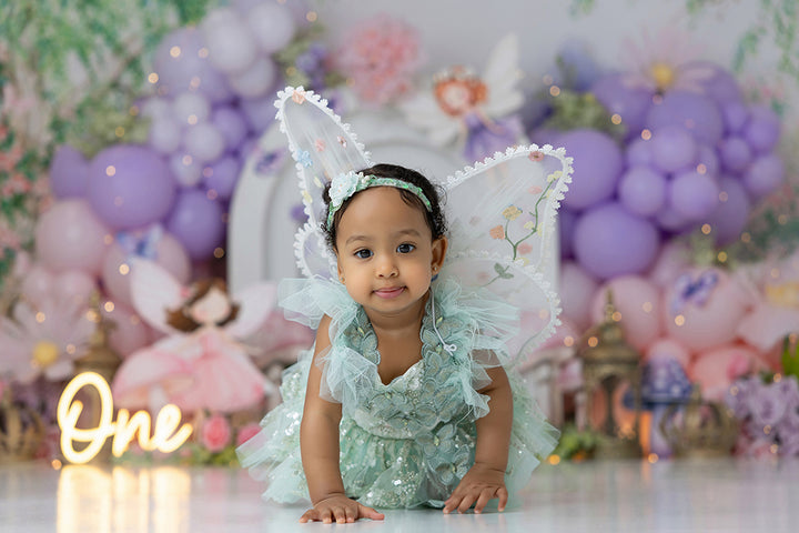 Fairies &amp; Flowers cake smash backdrop with pastel balloon garlands, fairy garden details, flowers, mushrooms, butterflies, lanterns, greenery, and enchanted birthday styling.