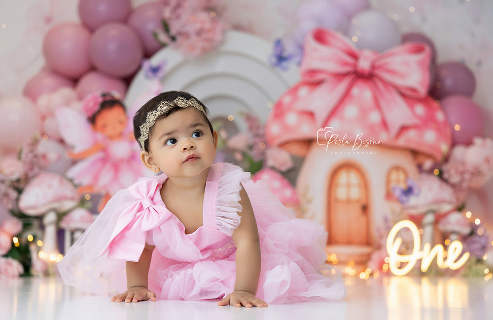 Where Fairies Bloom cake smash backdrop with pastel mushroom cottage, pink and lavender balloon garlands, oversized flowers, butterflies, lantern details, twinkle lights, and enchanted fairy garden styling.