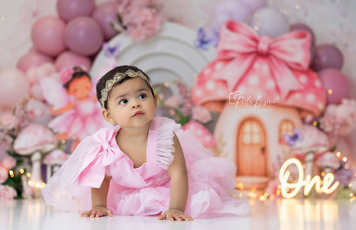 Where Fairies Bloom cake smash backdrop with pastel mushroom cottage, pink and lavender balloon garlands, oversized flowers, butterflies, lantern details, twinkle lights, and enchanted fairy garden styling.