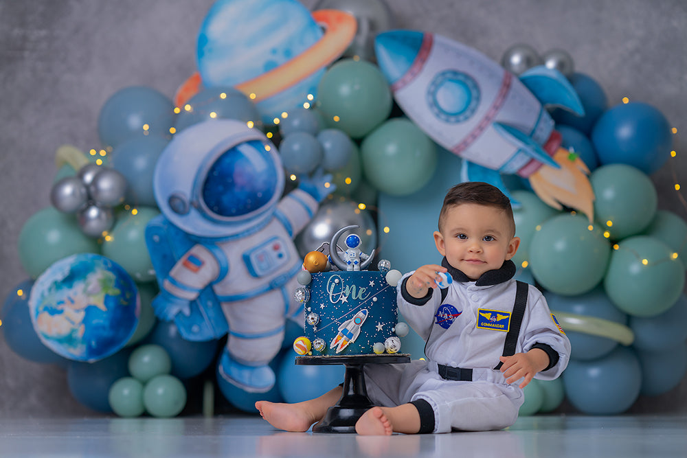 Galaxy Explorer cake smash backdrop with blue and teal balloon garlands, astronaut, rocket ship, planets, metallic accents, and outer space birthday styling.