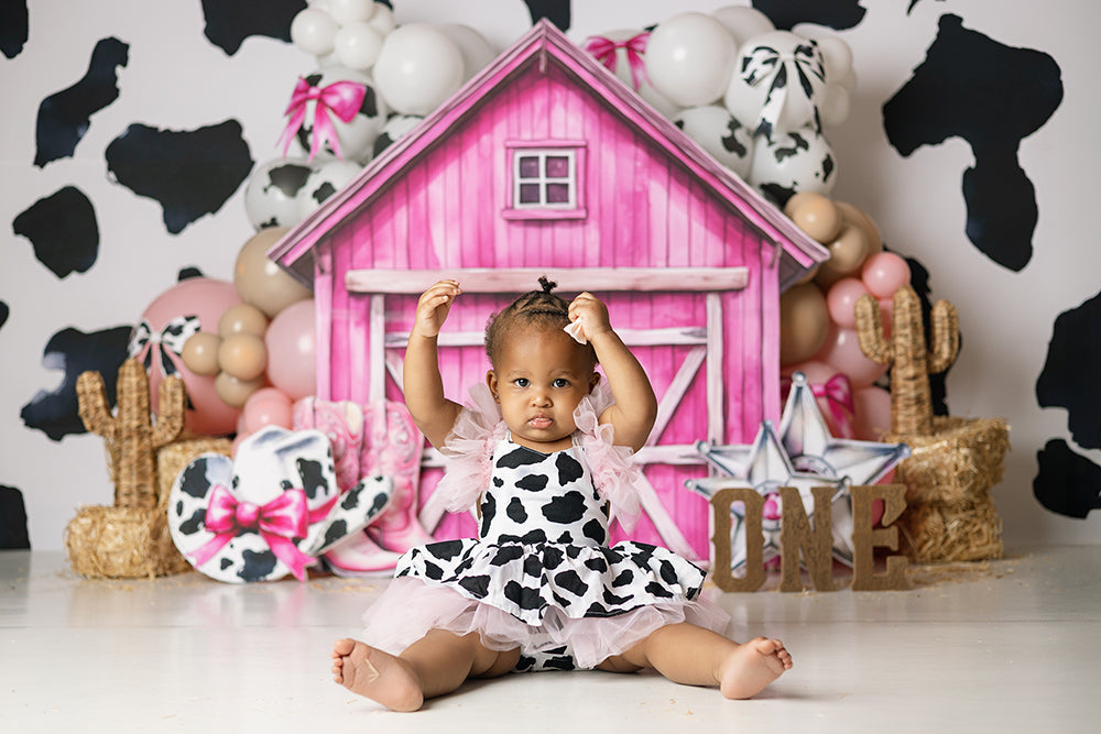 Pink Cowgirl Barn cake smash backdrop with bright pink barn, cow print wall, pastel balloon garlands, cowgirl boots, hay bales, sheriff star, rope cactus accents, and western birthday studio styling.