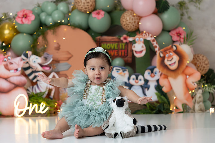 Move It! Move It! Jungle Animals photography backdrop featuring adorable lions, giraffes, zebras, and lemurs surrounded by sage green, blush pink, peach, ivory, and giraffe-print balloons, tropical greenery, pink hibiscus flowers, and a charming wooden safari hut for first birthday and cake smash portraits.