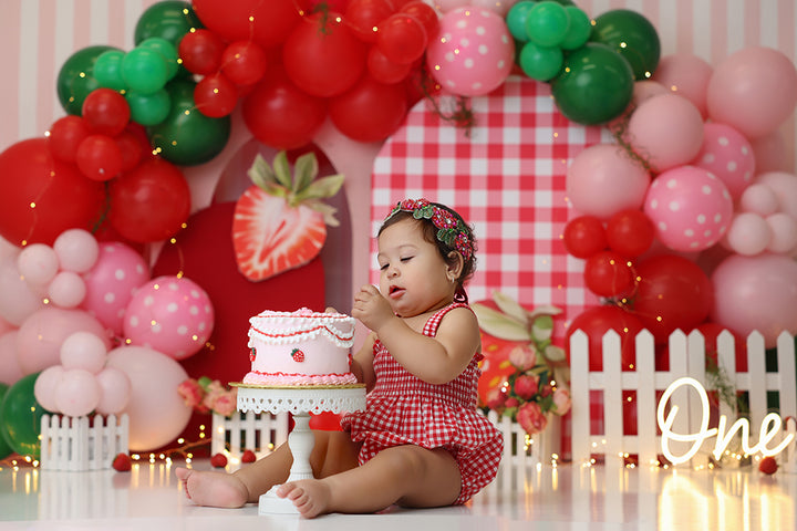 Strawberry-themed cake smash backdrop with balloon arch, gingham, and picket fence – Picked Just for You by HSD Backdrops.