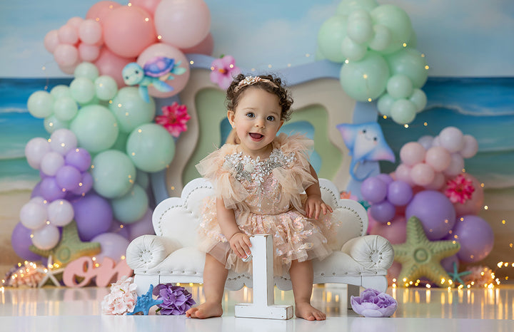 Pastel under-the-sea backdrop with balloons, sea animals, and coral on the beach.