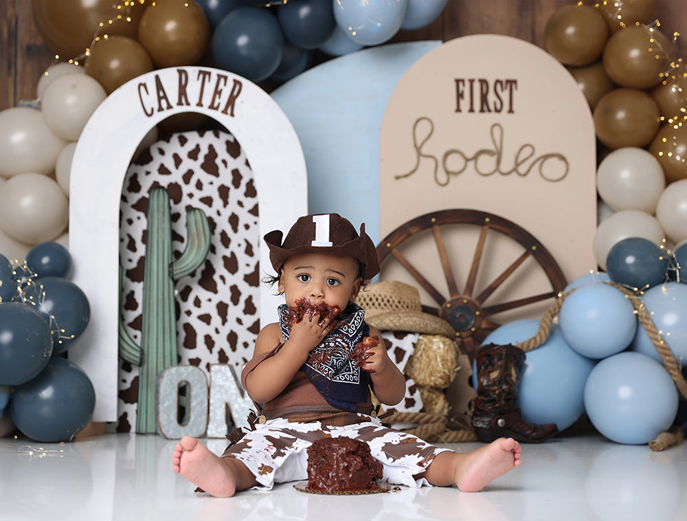 Western cowboy first birthday backdrop with cowhide, balloons, and wagon wheel props.
