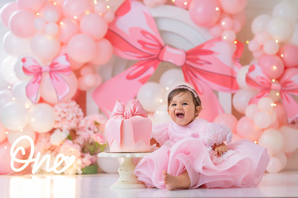 Pink and white balloon birthday backdrop with bow decorations and stacked faux cakes.
