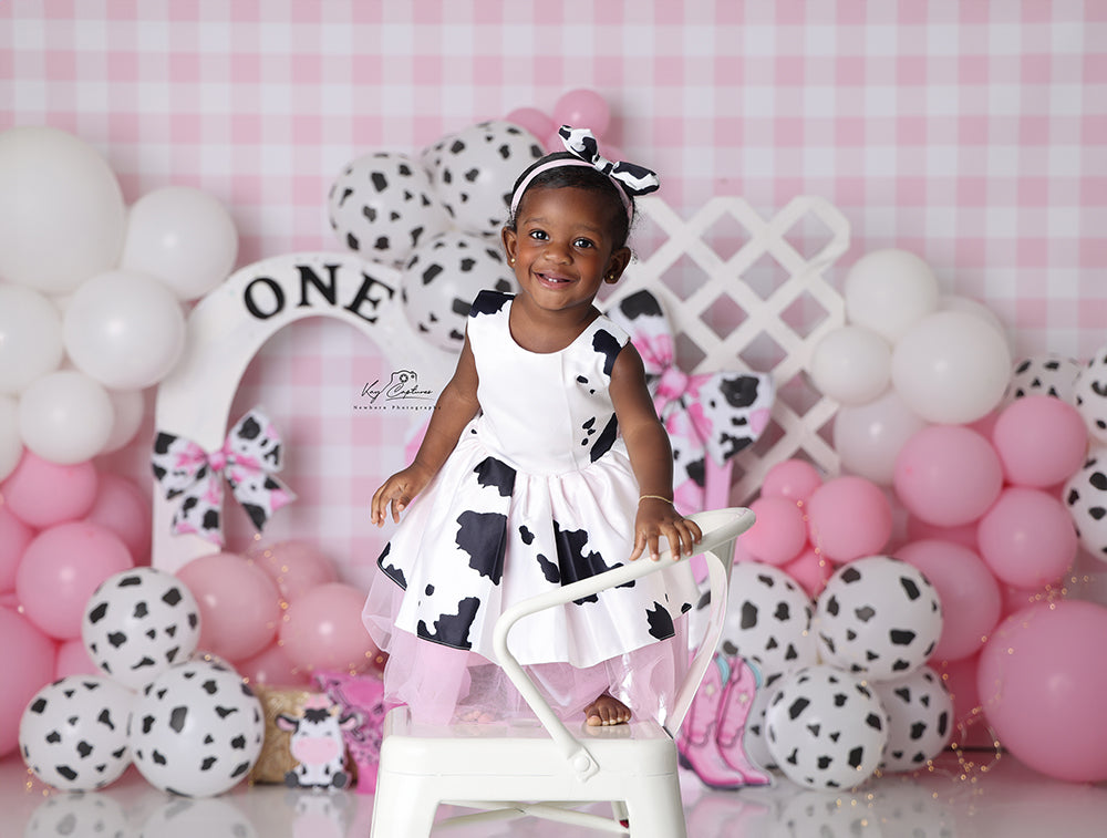 Pink cow print birthday backdrop with barn, bows, balloons, and cowboy boots for 1st birthday photos.