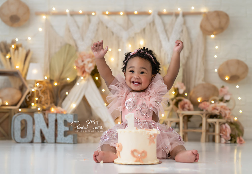 Boho One neutral macrame cake smash backdrop with pampas grass, warm lights, and floral decor for first birthday photoshoots.