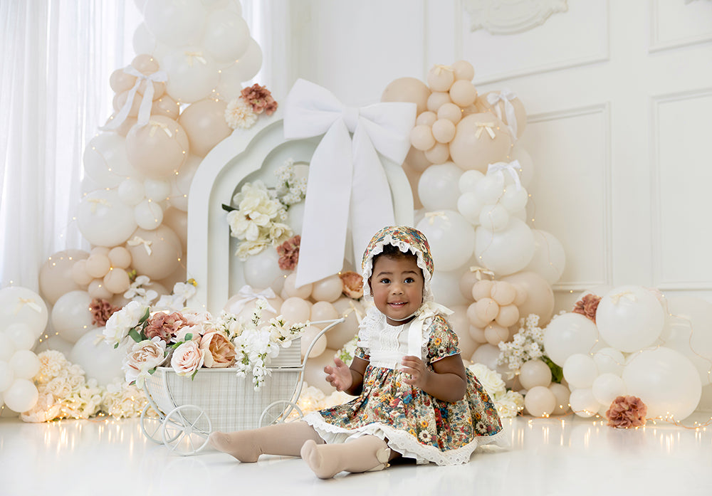 Ivory Bloom photography backdrop with white and beige balloons, large white bow, and floral accents for elegant cake smash and birthday photo sessions.