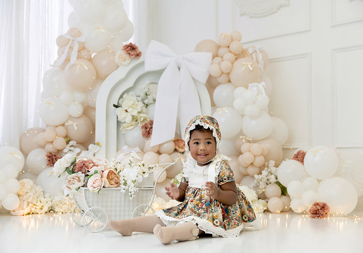 Ivory Bloom photography backdrop with white and beige balloons, large white bow, and floral accents for elegant cake smash and birthday photo sessions.