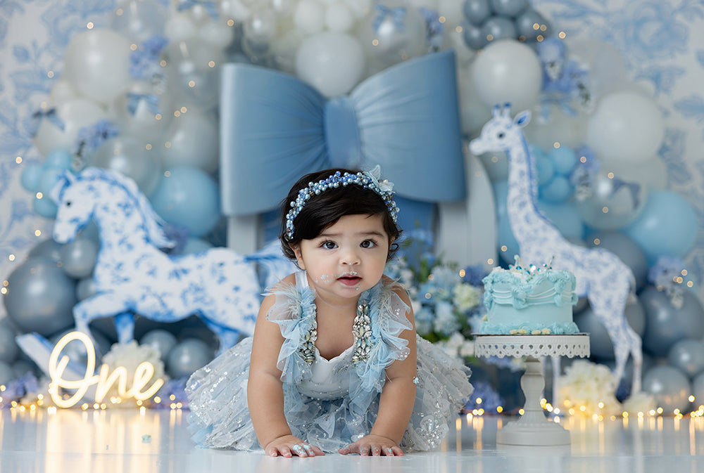 Blue toile safari cake smash backdrop with rocking horse, giraffe, balloons, and oversized blue bow.