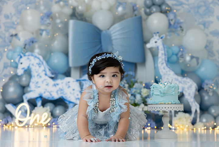 Blue toile safari cake smash backdrop with rocking horse, giraffe, balloons, and oversized blue bow.