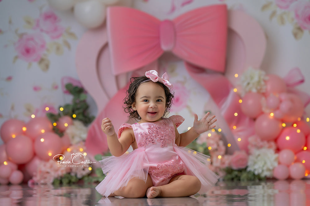 Blush pink bow with floral backdrop design featuring roses, hydrangeas, balloons, and greenery for a girly cake smash or milestone photo session.