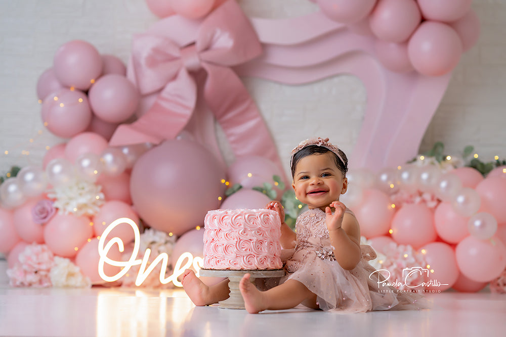 Pink bow birthday backdrop with blush balloons, pearl accents, and soft florals for girl’s cake smash and milestone photography.