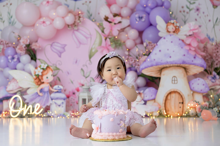 Pastel fairy garden cake smash photography backdrop with enchanted mushroom house and pink balloon clusters for girl first birthday sessions.