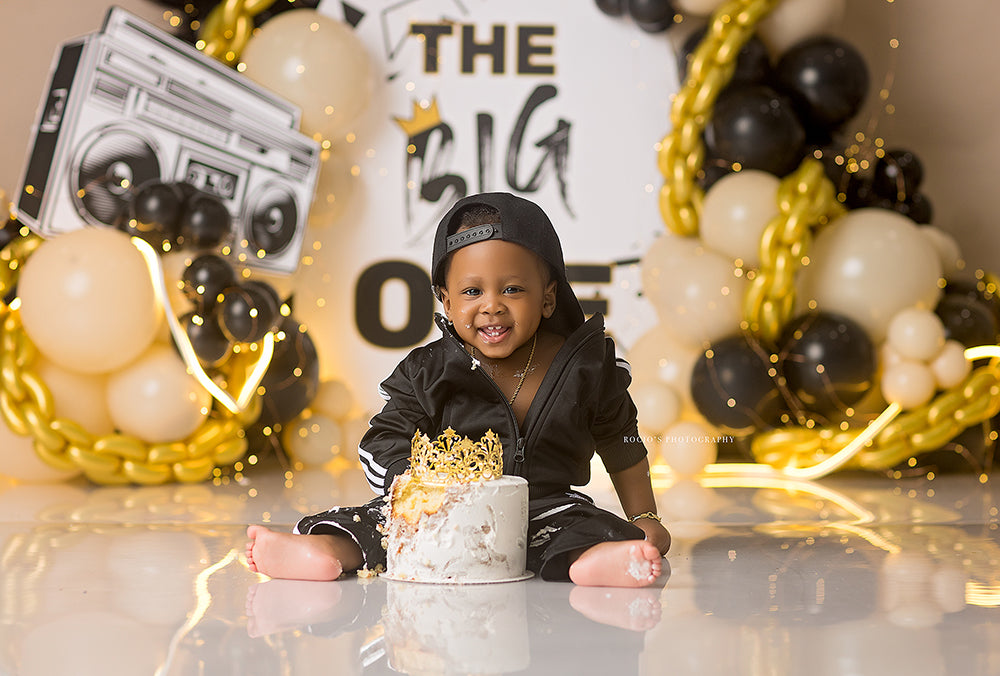The Big One boys cake smash photography backdrop with black and gold balloons, chain accents, crown detail, and bold first birthday styling.